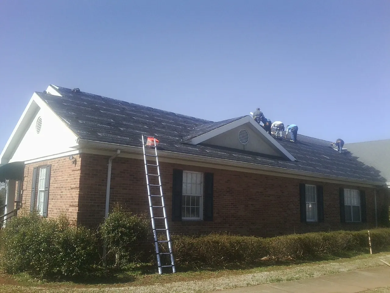 Crew tearing shingles off a brick ranch roof with a ladder set — Emergency Roof Repair work by our crew in Churdan