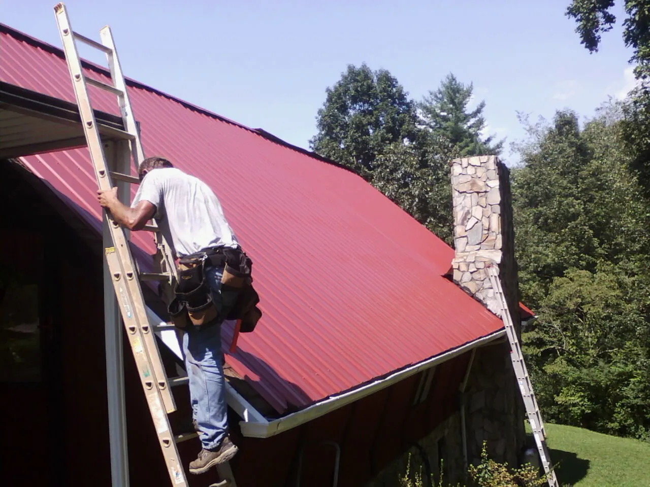 Roofer on a ladder beside a red metal roof and stone chimney — Soffit & Fascia Repair by Roofing Brothers in Churdan